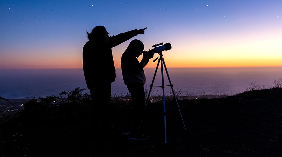 Silhouette of two figures looking through a telescope at dusk 